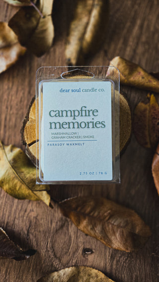 A packaged wax melt labeled "campfire memories" by Dear Soul Candle Co. sits on a rustic wooden surface surrounded by dried autumn leaves.
