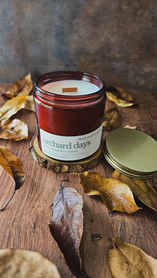 A red candle jar labeled 'Orchard Days' with a wood wick, styled on a wooden surface with scattered dried autumn leaves and a gold-toned metal lid beside it.